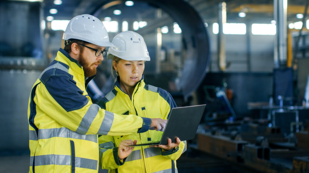 engineers in hardhats and high-visibility jackets reviewing a laptop in a factory.