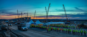 construction workers gathering at dusk for an infrastructure hiring surge event under cranes.